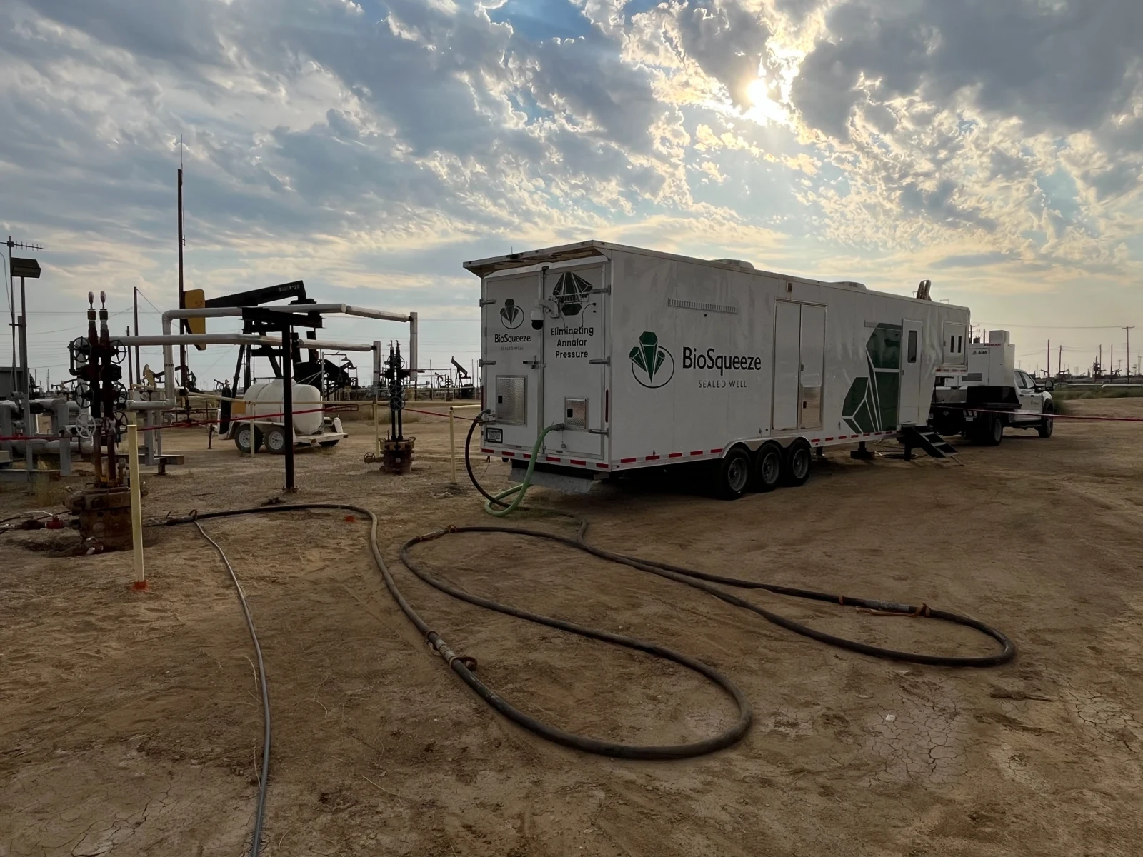 BioSqueeze treatment trailer beside surface well equipment beneath a sunlit cloudy sky.